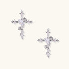 Pair of ornate silver cross earrings with heart-cut clear zirconia center, rose motifs, and spike tips on cream background.